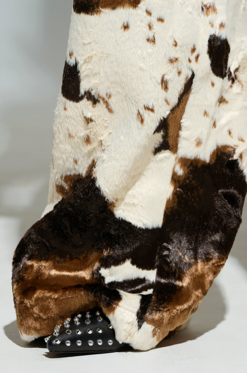 A close-up of a fur coat with cow-print detailing, draped over spiky, studded high-heeled shoes that perfectly match the FEELING IT COW TROUSER. The coat showcases a blend of brown, white, and black patches.