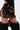 A person wearing the DONT THINK ABOUT IT COW PRINT MINI SKIRT IN BROWN with lace-up sides, black furry leg warmers, and a shiny black top. Only the lower half of the outfit is visible.