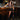 A woman stands in front of a bar, wearing the DONT THINK ABOUT IT COW PRINT MINI SKIRT IN BROWN with a sleeveless vest and tall black fringe boots. The bar features wooden chairs, glasses, beer taps, and a "BELIEVE" sign in the background.