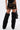 Someone wears the DONT THINK ABOUT IT COW PRINT MINI SKIRT IN BROWN with tall black furry leg warmers over black shoes, holding a black handbag. The image is cropped from the waist down on a plain white background.