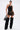 A woman poses confidently in a black halter top, the DONT THINK ABOUT IT COW PRINT MINI SKIRT IN BROWN, black furry leg warmers, pointed lace-up boots, and holding a black handbag against a plain white background.
