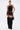 A woman models the DONT THINK ABOUT IT COW PRINT MINI SKIRT IN BROWN with a black halter top, tall black furry boots, large hoop earrings, and a black handbag against a plain white background.