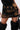 A person wearing the DONT THINK ABOUT IT COW PRINT MINI SKIRT IN BROWN, a black top, black faux fur leg warmers, and holding a black handbag against a plain background.