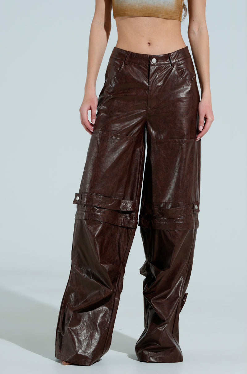 A person is wearing the TOO GOOD FAUX LEATHER WIDE LEG TROUSER in dark brown, paired with a crop top that reveals part of the midriff. The setting features a plain, light-colored background.
