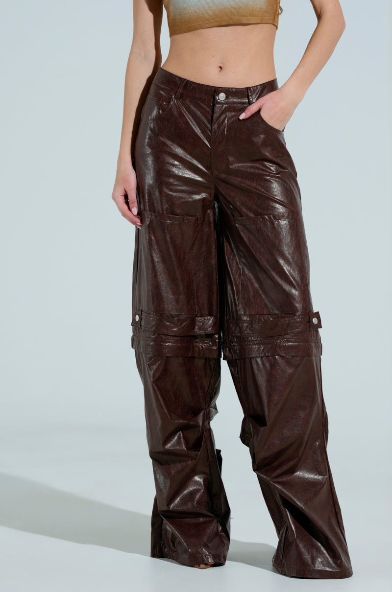 Sporting TOO GOOD FAUX LEATHER WIDE LEG TROUSERS in shiny brown, someone showcases their midriff with a relaxed fit and spacious pockets against a plain light gray background.