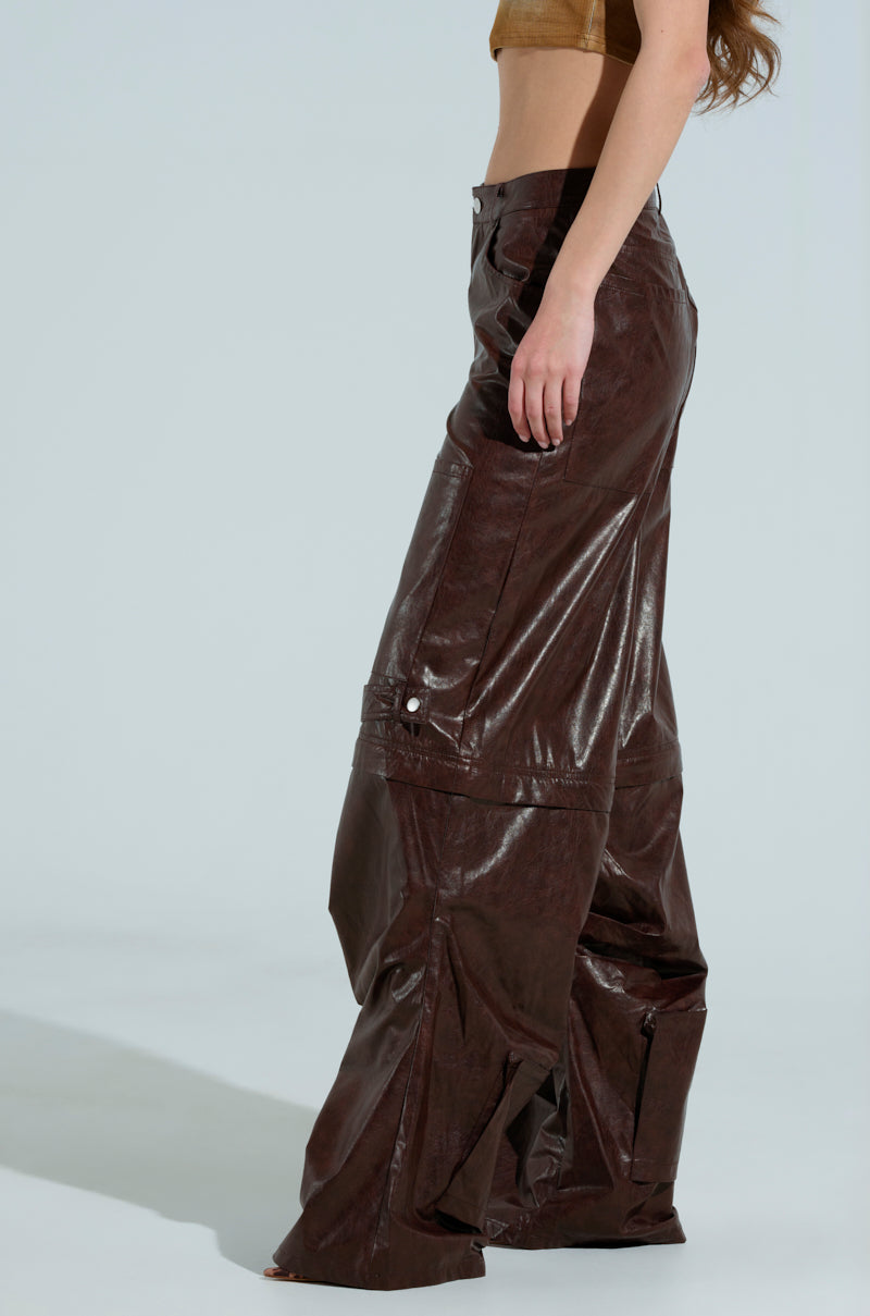 Side view of a person wearing a sleeveless brown top paired with the TOO GOOD FAUX LEATHER WIDE LEG TROUSER, reflecting light on a plain light gray background. The person's face is not visible.