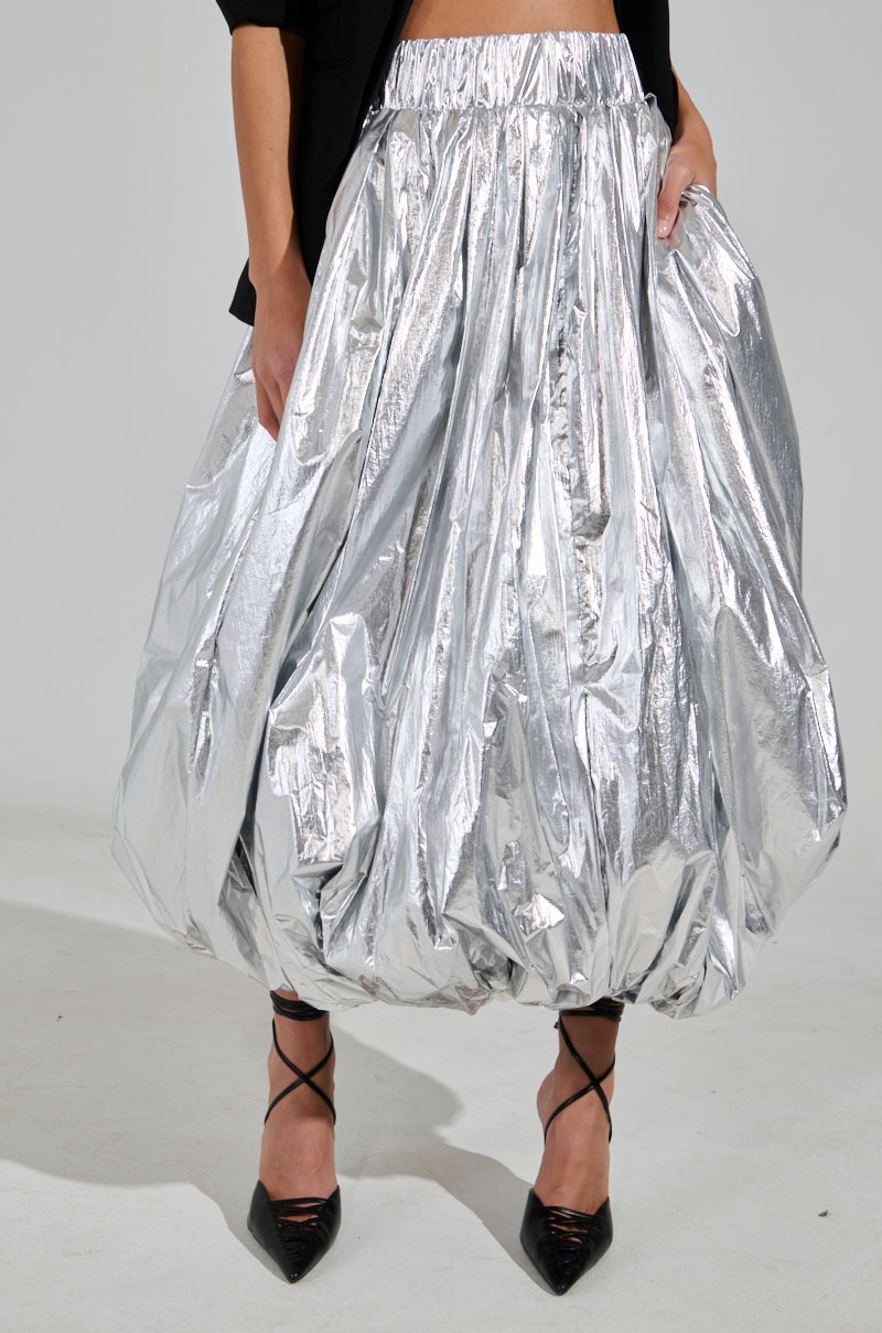 Wearing the MIDNIGHT LOVERS MAXI BUBBLE SKIRT, a shiny metallic bubble-style with an elastic waistband, paired with black pointed heels and a black top offers a bold, stylish look.