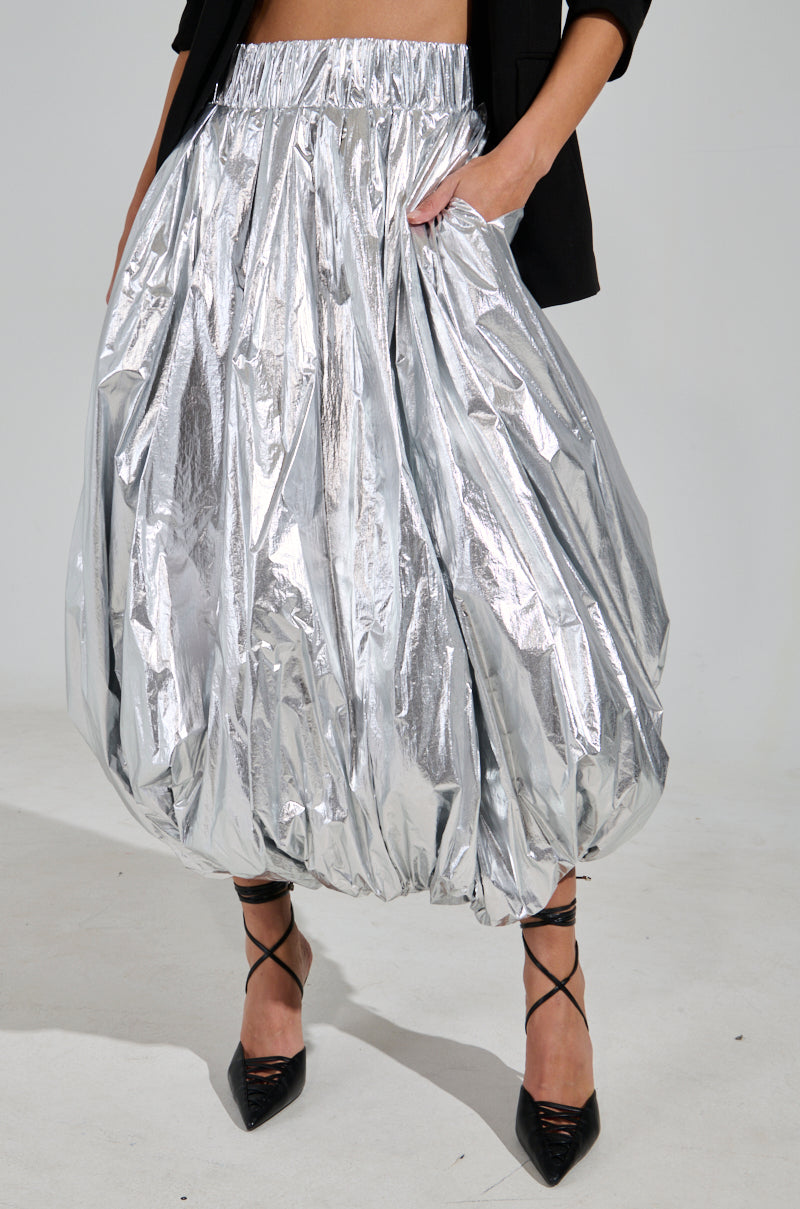 A person in the MIDNIGHT LOVERS MAXI BUBBLE SKIRT, showcasing its shiny silver metallic fabric and loose, gathered fit, paired with black pointed heels. They also don a black blazer with one hand casually in their pocket.