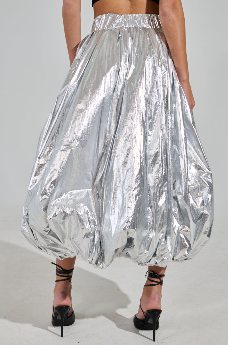 A person in the MIDNIGHT LOVERS MAXI BUBBLE SKIRT, featuring a metallic fabrication with a balloon silhouette, stands on a light-colored floor against a plain backdrop. Paired with black high heels, the skirt creates a shimmering effect.