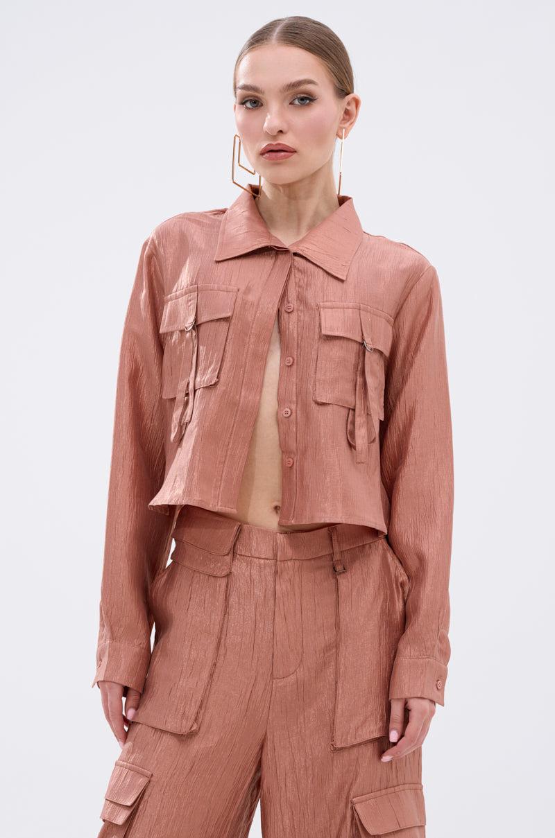 A woman wears the NOT ALONE BUTTON DOWN TOP from AKIRA Label, a shiny light brown metallic blouse, paired with matching high-waisted cargo pants and posing against a plain white background.