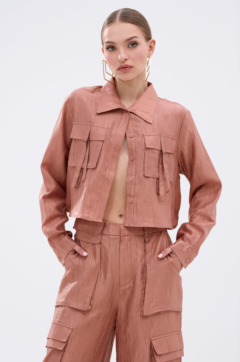 A woman wears the NOT ALONE BUTTON DOWN TOP in dusty pink, featuring a cropped boxy fit and flap pockets, paired with matching high-waisted pants, standing with her hands in her pockets against a plain light background.