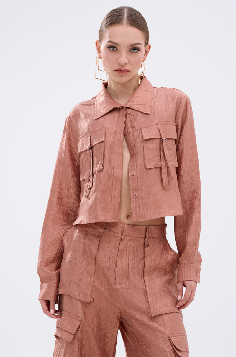 A woman models the NOT ALONE BUTTON DOWN TOP in a shiny, textured rose-gold finish with front pockets and matching cargo pants, styled with slicked-back hair and bold geometric earrings against a plain background.