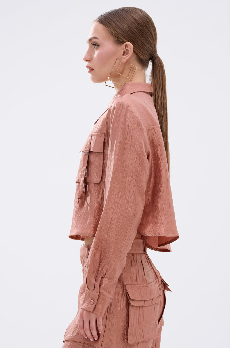 A woman with straight brown hair in a low ponytail wears AKIRA Label’s NOT ALONE BUTTON DOWN TOP in textured dusty pink with matching high-waisted pants, styled with large geometric earrings as she stands in profile.