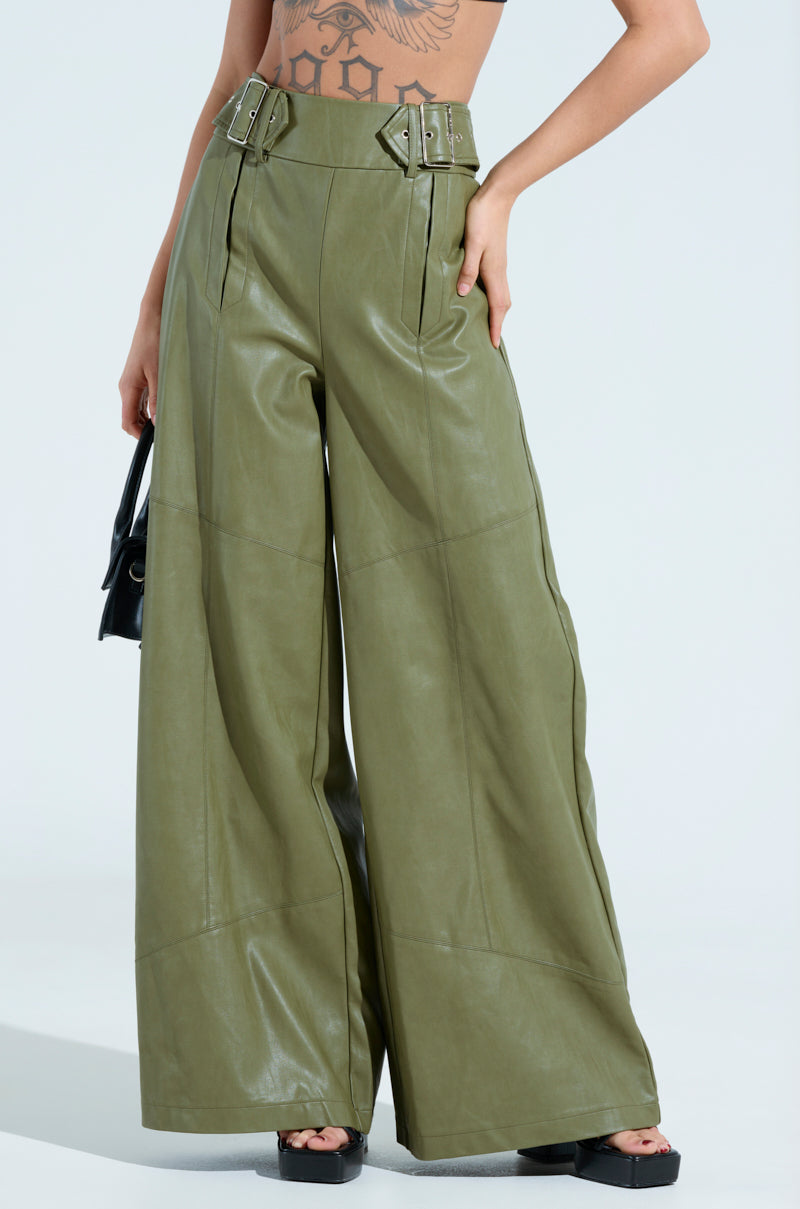A person wearing the AMY TROUSER IN OLIVE featuring a high-rise design, faux leather wide-leg style, and waist belt buckles pairs it with black platform sandals and holds a small black purse against a light blue background.