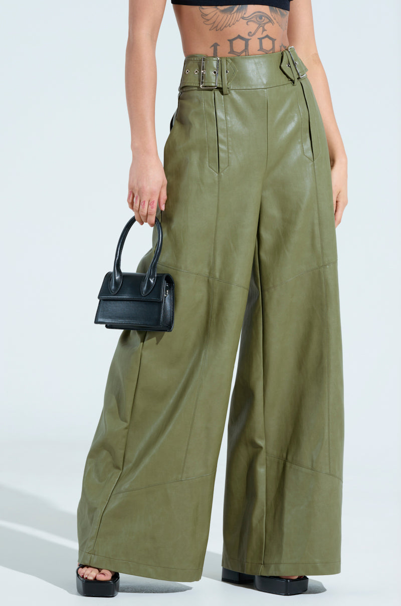 Wearing the AMY TROUSER IN OLIVE, which features high-rise faux leather and a chic wide-leg silhouette with belt details, a person holds a small black handbag. A tattoo peeks from their torso in a shot captured from midsection to floor.