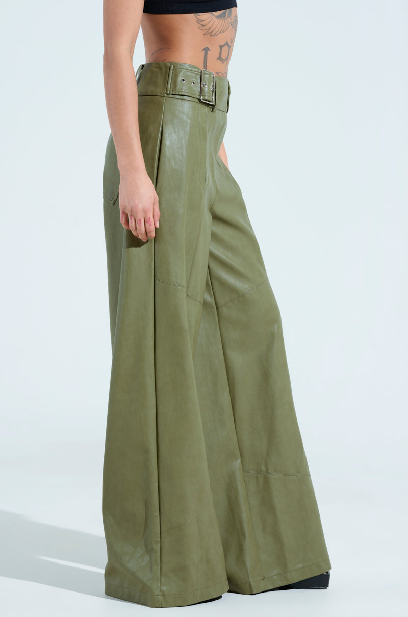 Someone dressed in the AMY Trouser in olive, featuring wide-leg and high-rise design with a matching belt, pairs it with a black crop top that reveals a tattoo on their left side. The scene is set against a plain white backdrop.