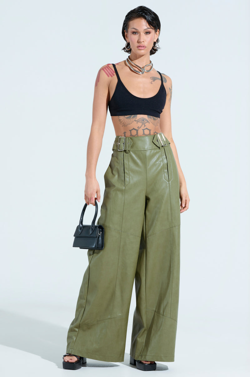 A person with short hair models black crop top and AMY TROUSER IN OLIVE, emphasizing the wide-leg silhouette. They carry a small black handbag, revealing tattoos on their arms and torso, paired with black platform sandals.