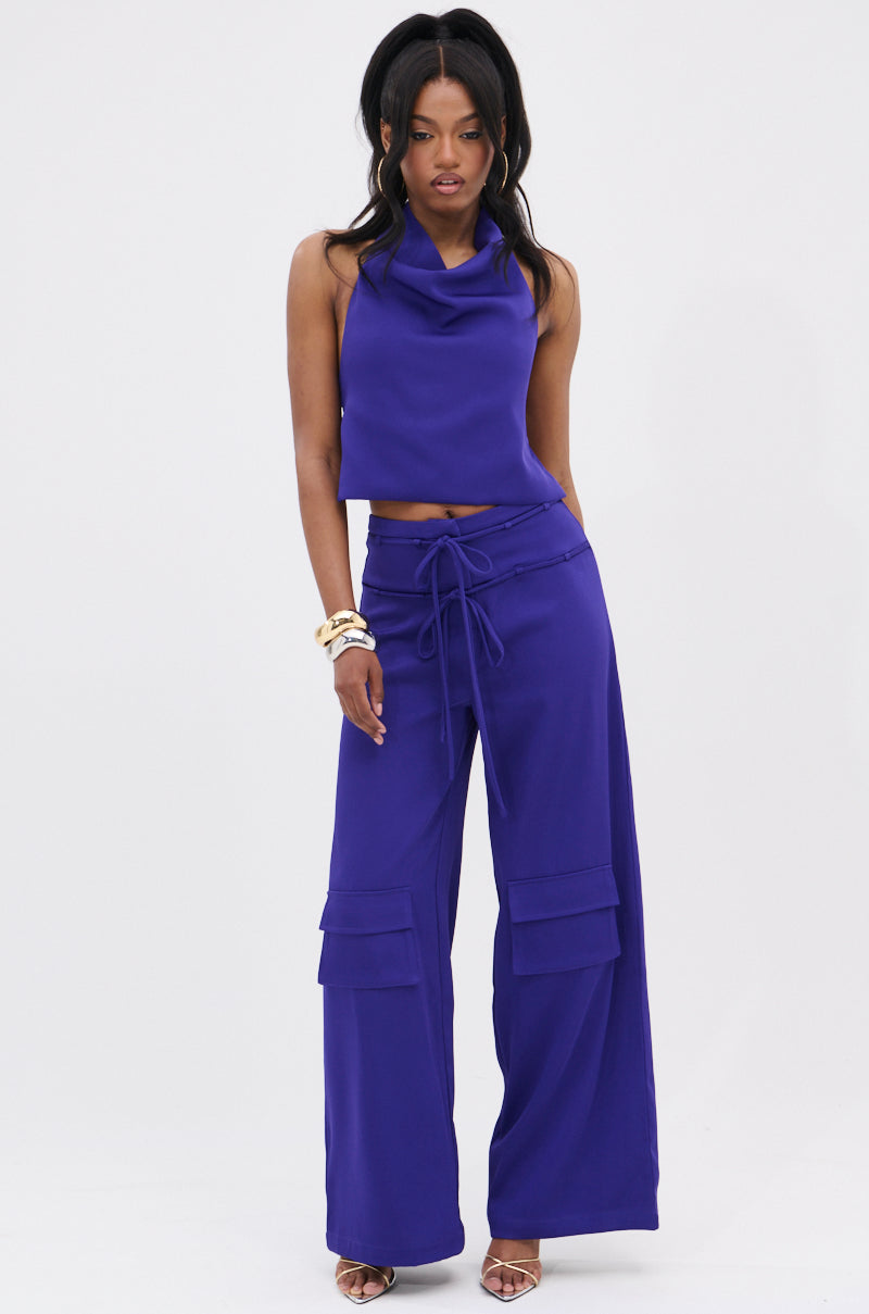 A woman poses against a white background wearing the NEEDED THIS HALTER TOP in sleeveless cobalt blue with an open back, paired with matching wide-leg cargo pants with a drawstring waist. She completes the look with gold bracelets and strappy heels.
