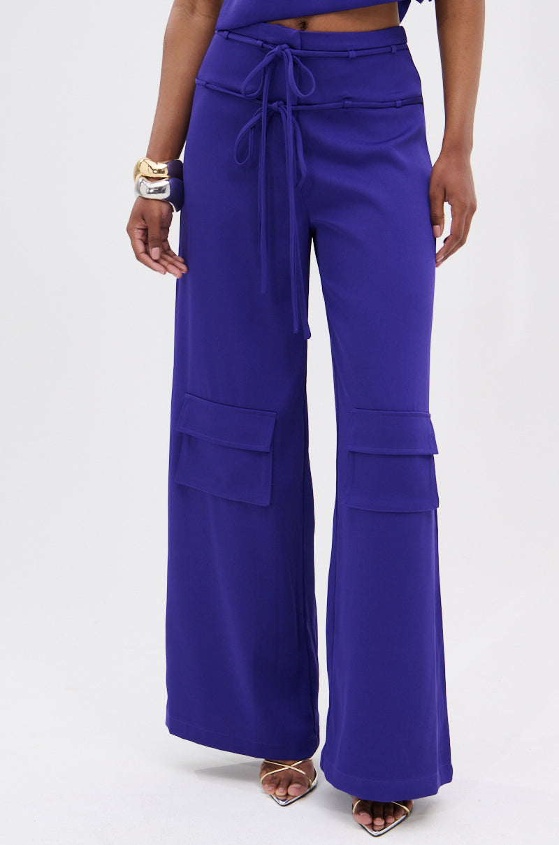 A person wears the NEEDED THIS CARGO TROUSER: vibrant purple, high-rise, wide-leg pants featuring front pockets and a tie waist, styled with gold heels and a chunky gold bracelet against a plain light background.