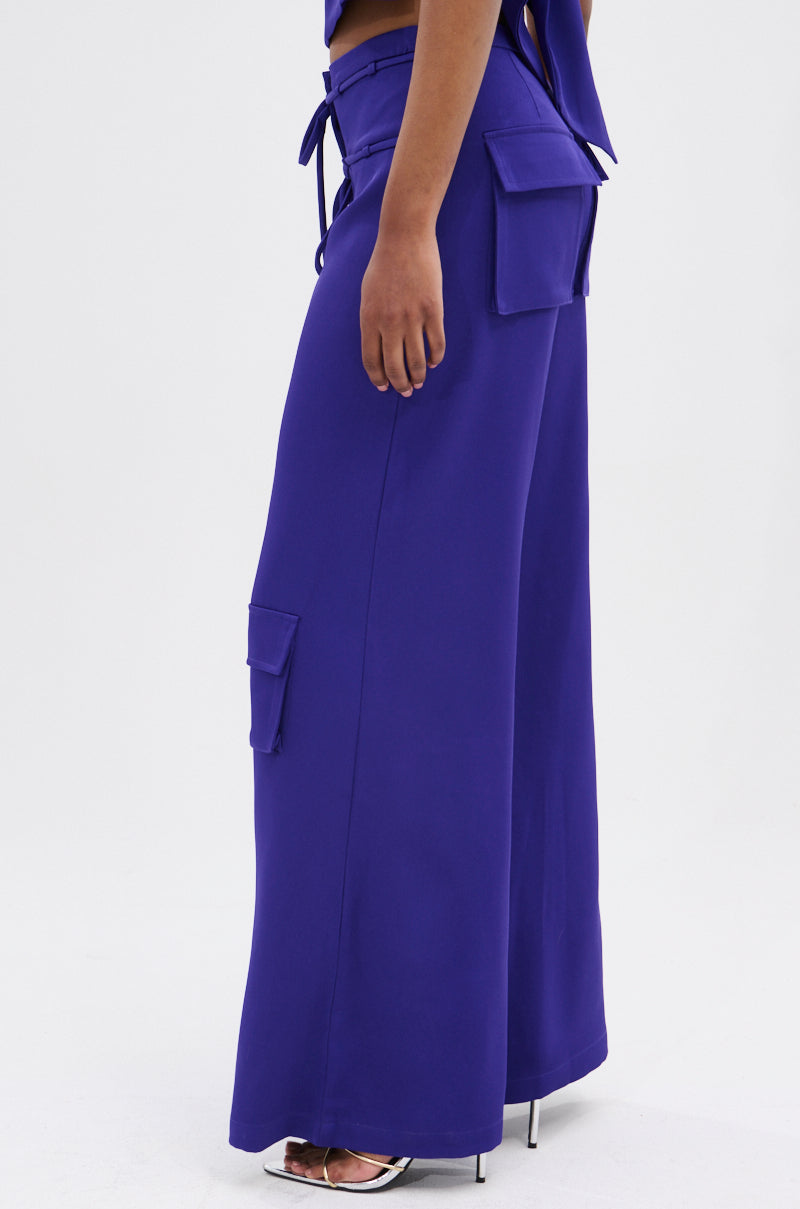 A person models the NEEDED THIS CARGO TROUSER in vibrant purple high-rise wide-leg style with large side pockets, paired with silver high-heeled sandals, shown from the side against a plain white background.