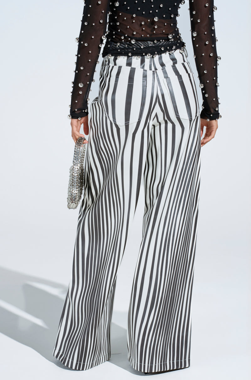 A person is wearing high-rise, black-and-white vertically striped PARADOX STRIPE PANT and a black sheer top adorned with pearls. They hold a sparkling silver handbag against a plain, light-colored background.
