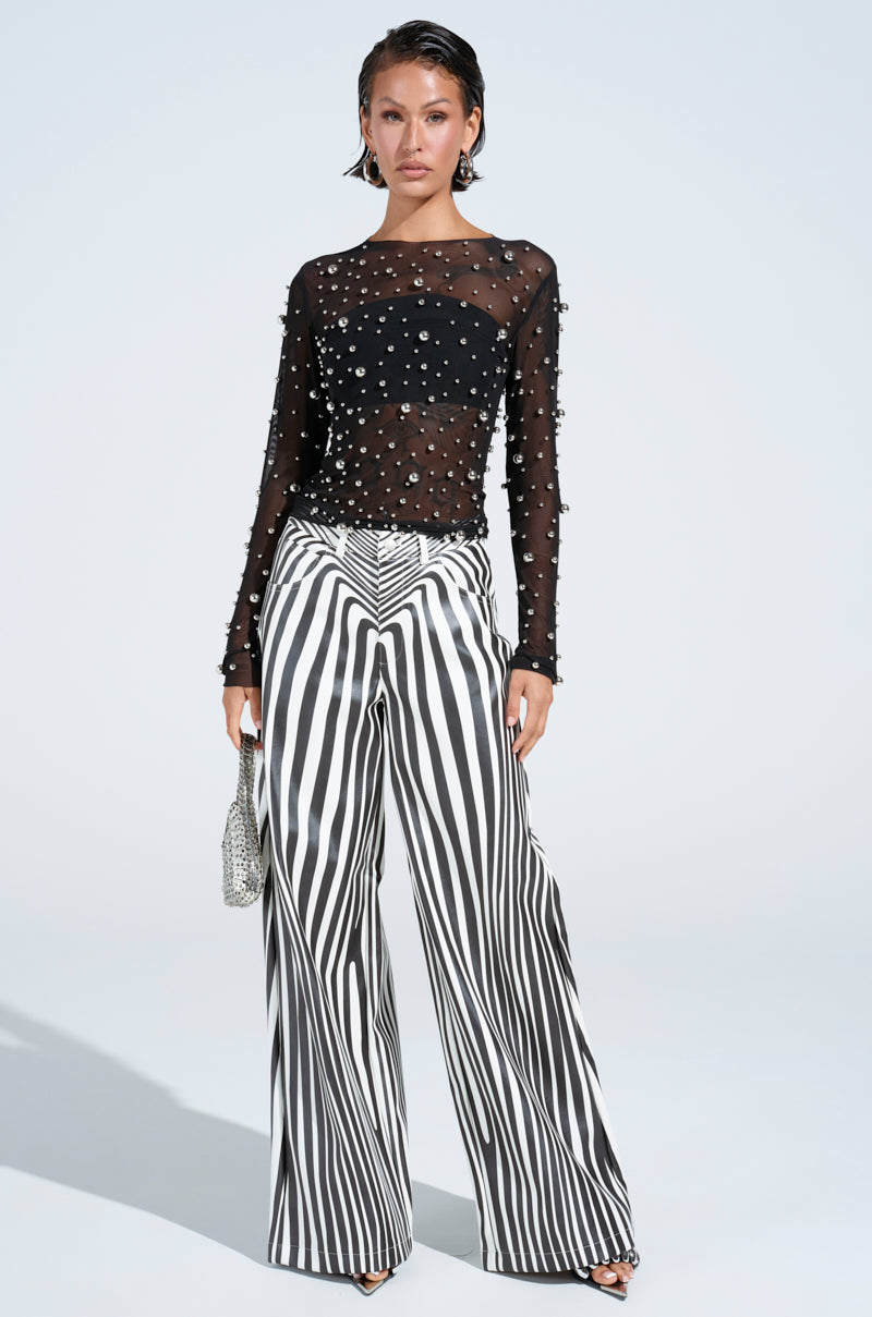 A woman poses confidently against a white backdrop in a black sheer pearl-adorned top and chic PARADOX STRIPE PANT with a black-and-white design, holding a silver clutch.
