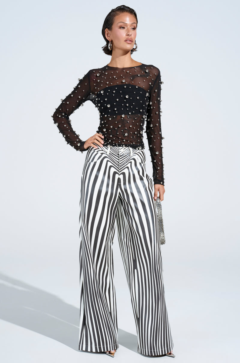 Dressed in PARADOX STRIPE PANTS with a high-rise fit and a sheer black pearl-adorned top, a person poses confidently, holding a small silver clutch. Their hair is styled back, and makeup highlights their features against a light background.