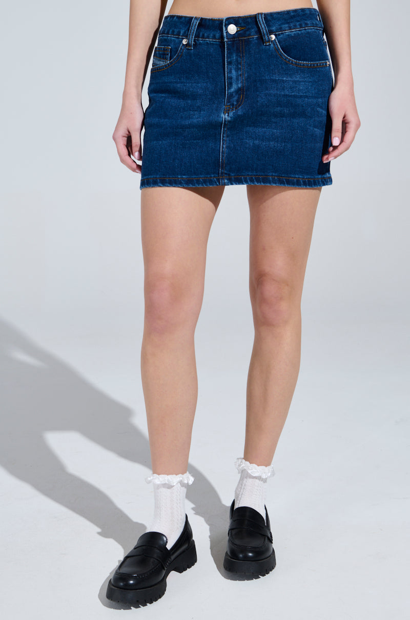 A person in a JOLENE DENIM MINI SKIRT with a high-rise fit, white ruffle socks, and black loafers stands against a plain gray background.