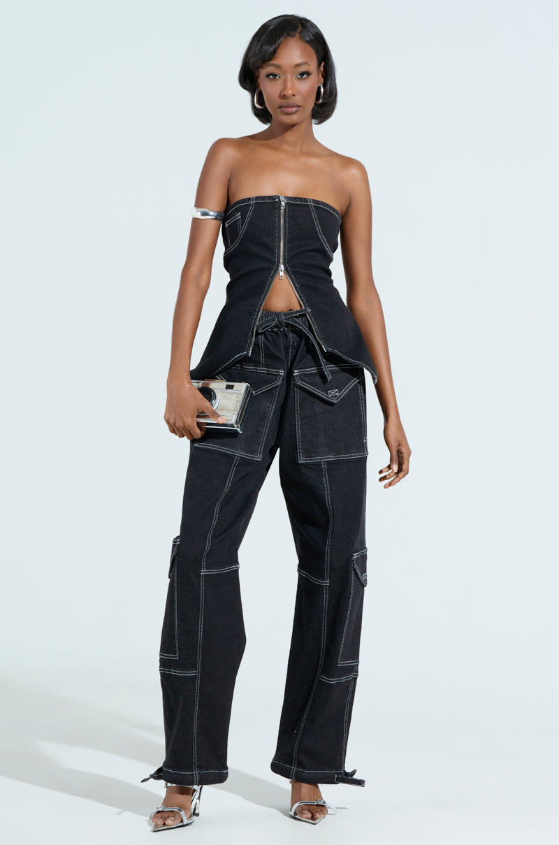 A woman exudes confidence in the BOUNDLESS CINCH PANT, a black, high-rise wide-leg design with white stitching and a matching strapless zipper front top. She accessorizes with a small purse and open-toe heels. Her sleek shoulder-length hair stands out against a light gray background.