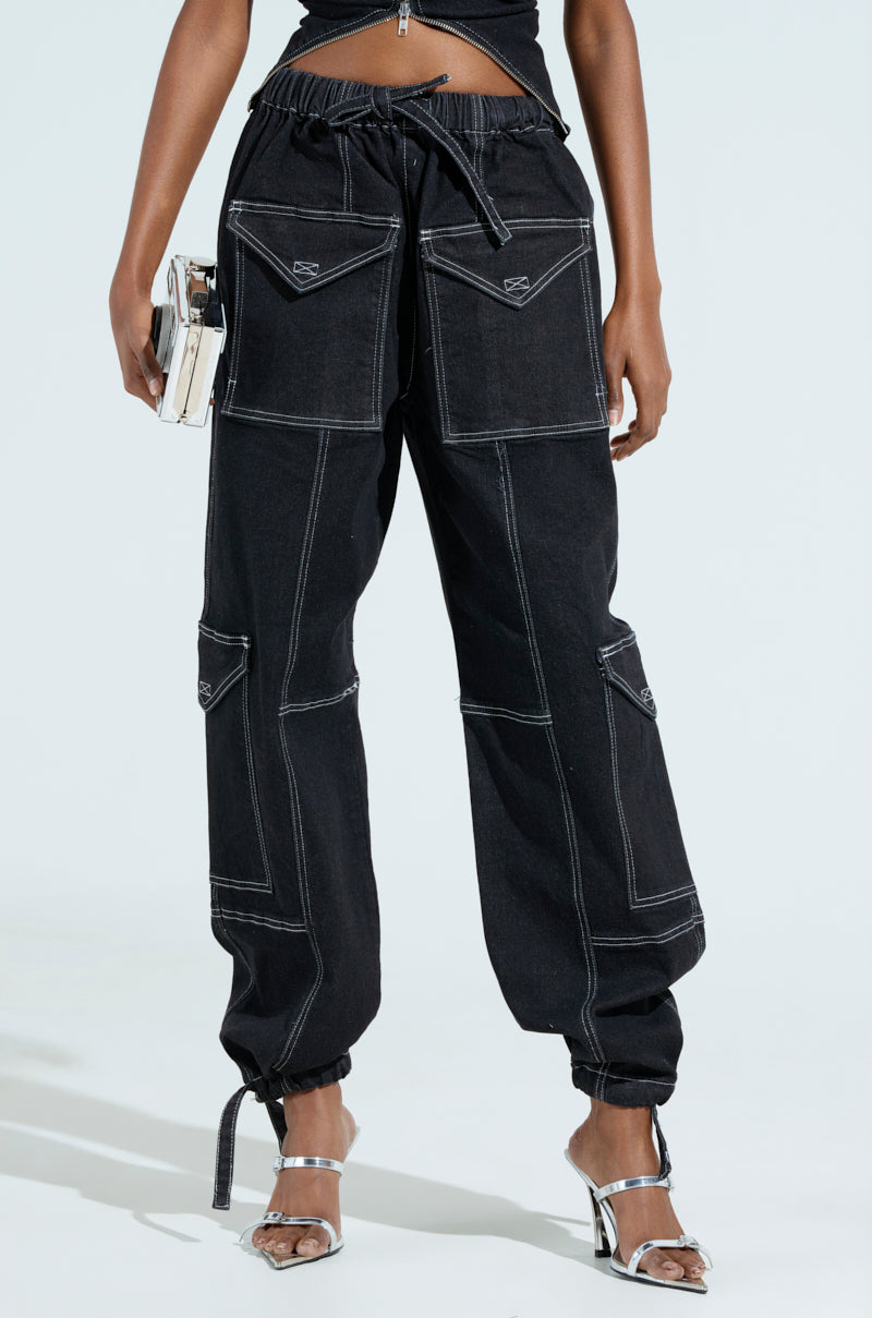 A person models the BOUNDLESS CINCH PANT, black with white stitching and a high-rise wide-leg design with large pockets. They hold a phone and wear silver strappy high-heeled sandals against a plain light-colored backdrop, enhancing the ensemble's chic appeal.