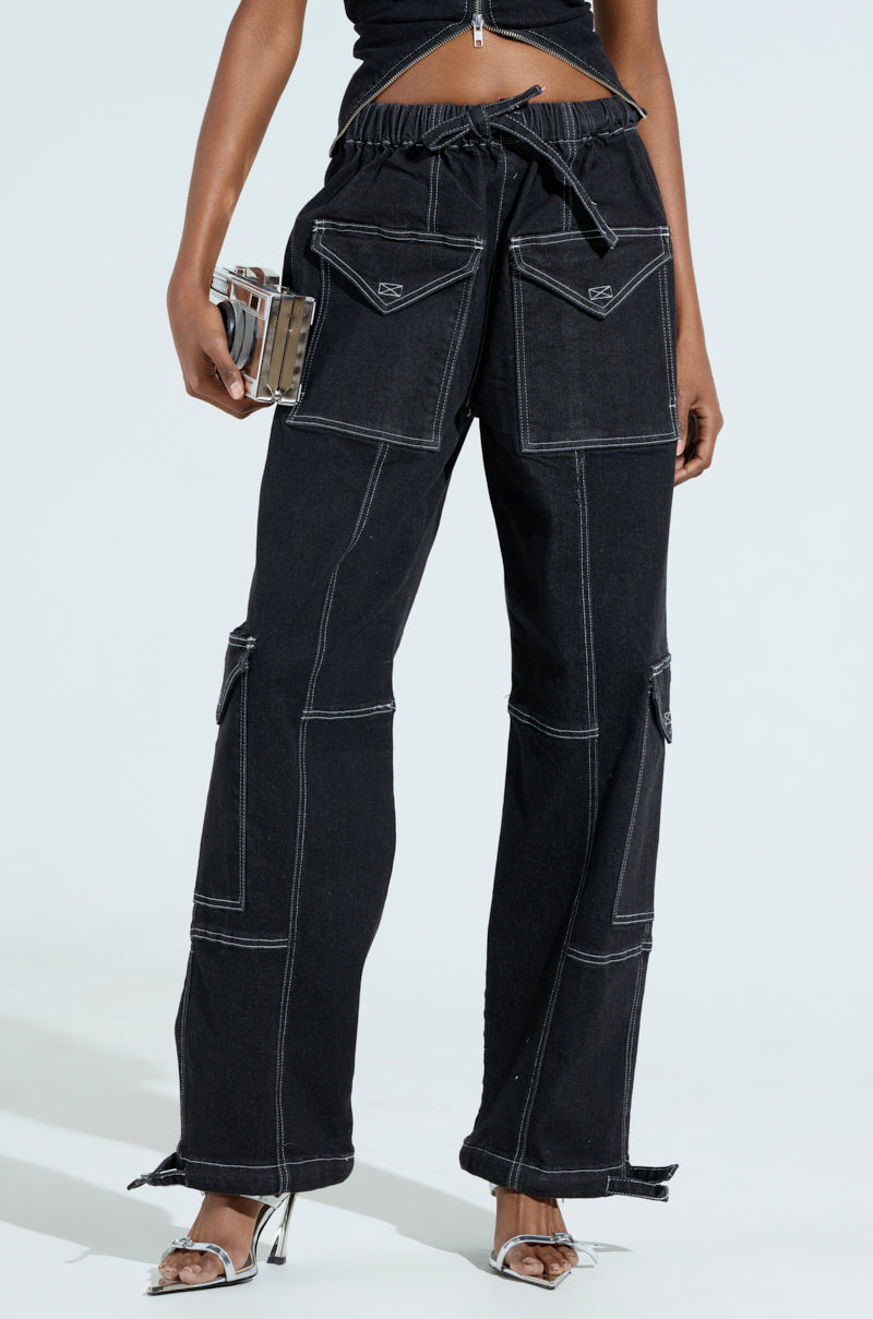 Wearing BOUNDLESS CINCH PANT, a person in black wide-leg jeans with white stitching pairs them with a matching top. They hold a small metallic handbag and wear silver high-heeled sandals against a plain, light-colored background.