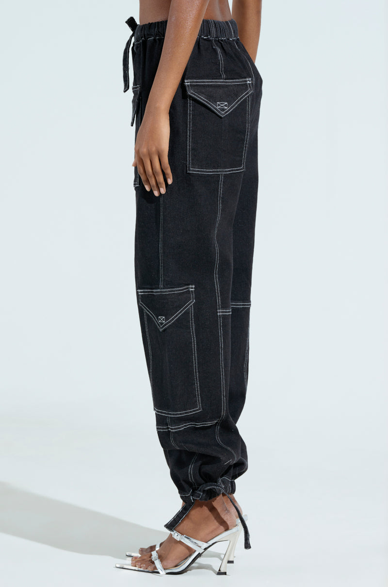 The BOUNDLESS CINCH PANT in black, with white stitching and large pockets, is showcased from the side. The design pairs elegantly with silver high-heeled sandals. Featuring drawstrings at the waist and ankles, these pants are striking against a plain white background.
