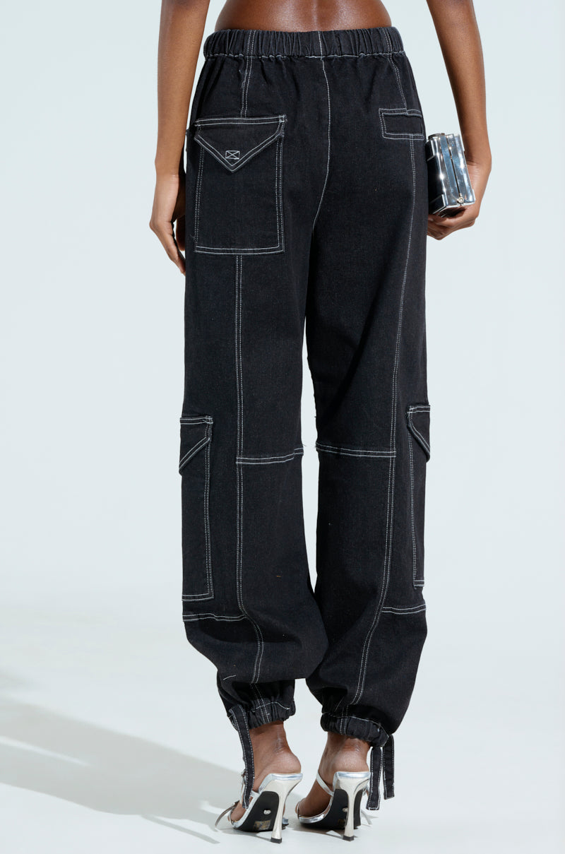 Someone is wearing the BOUNDLESS CINCH PANT, featuring a high-rise wide-leg design, black fabric with white stitching, large pockets, and elasticized cuffs. They're holding a metallic clutch and wearing high heels with transparent straps against a plain light-colored background.