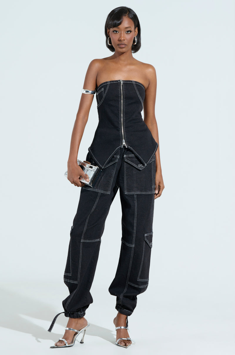 The person stands confidently in the black BOUNDLESS CINCH PANT, featuring defined white stitching and a high-rise wide-leg silhouette with a zipper front. Holding a clear clutch and wearing metallic high heels, they pose against a plain backdrop.
