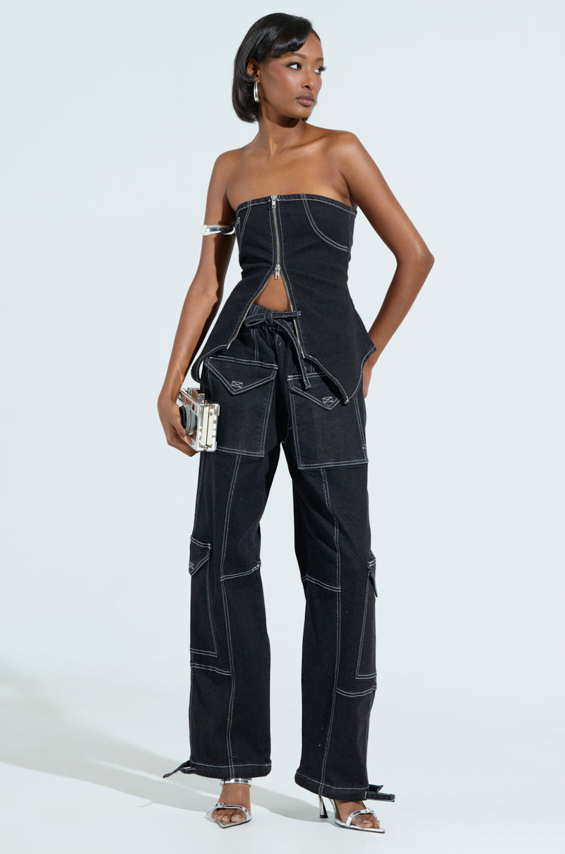 A person in a black denim strapless outfit, featuring BOUNDLESS CINCH PANT with high-rise wide-leg silhouette and a top with front zipper and white stitching, stands against a light background. They carry a clutch and wear silver heels, completing the chic look.