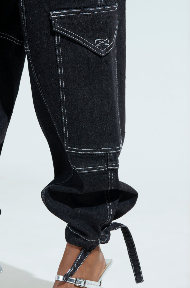 Close-up of black BOUNDLESS CINCH PANT with white stitching, showcasing a side flap pocket and strap. Paired with silver high-heeled sandals, these high-rise wide-leg pants create a chic contrast.
