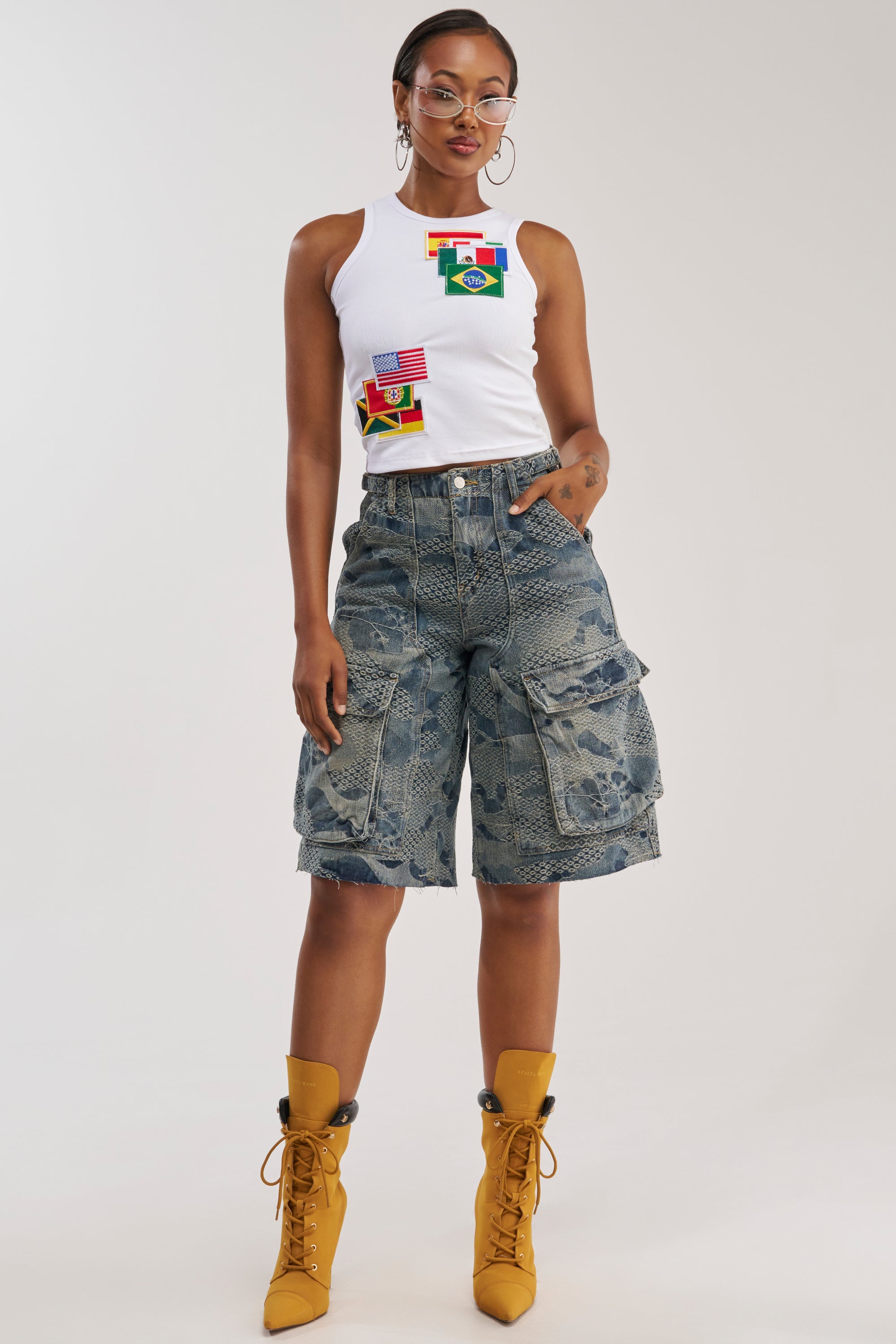 A woman stands facing forward against a plain background, wearing a white tank top with flag patches, large hoop earrings, yellow lace-up boots, and the I THINK YOU WANNA KNOW DENIM CARGO BERMUDA blue streetwear shorts.