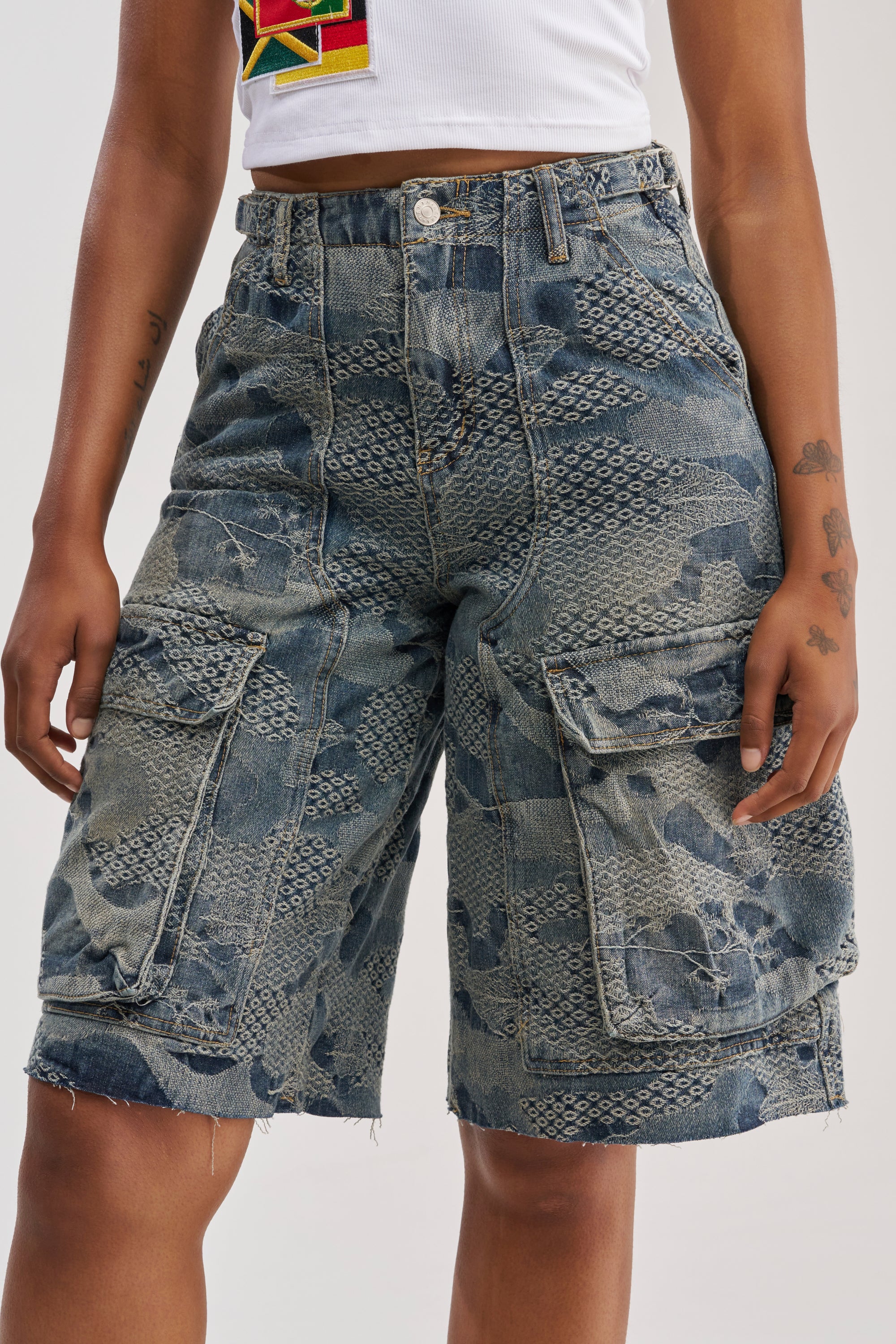 Wearing the I THINK YOU WANNA KNOW DENIM CARGO BERMUDA—patterned denim cargo bermuda shorts with frayed hems and large pockets—and a white crop top, this person shows off tattoos on their arms.