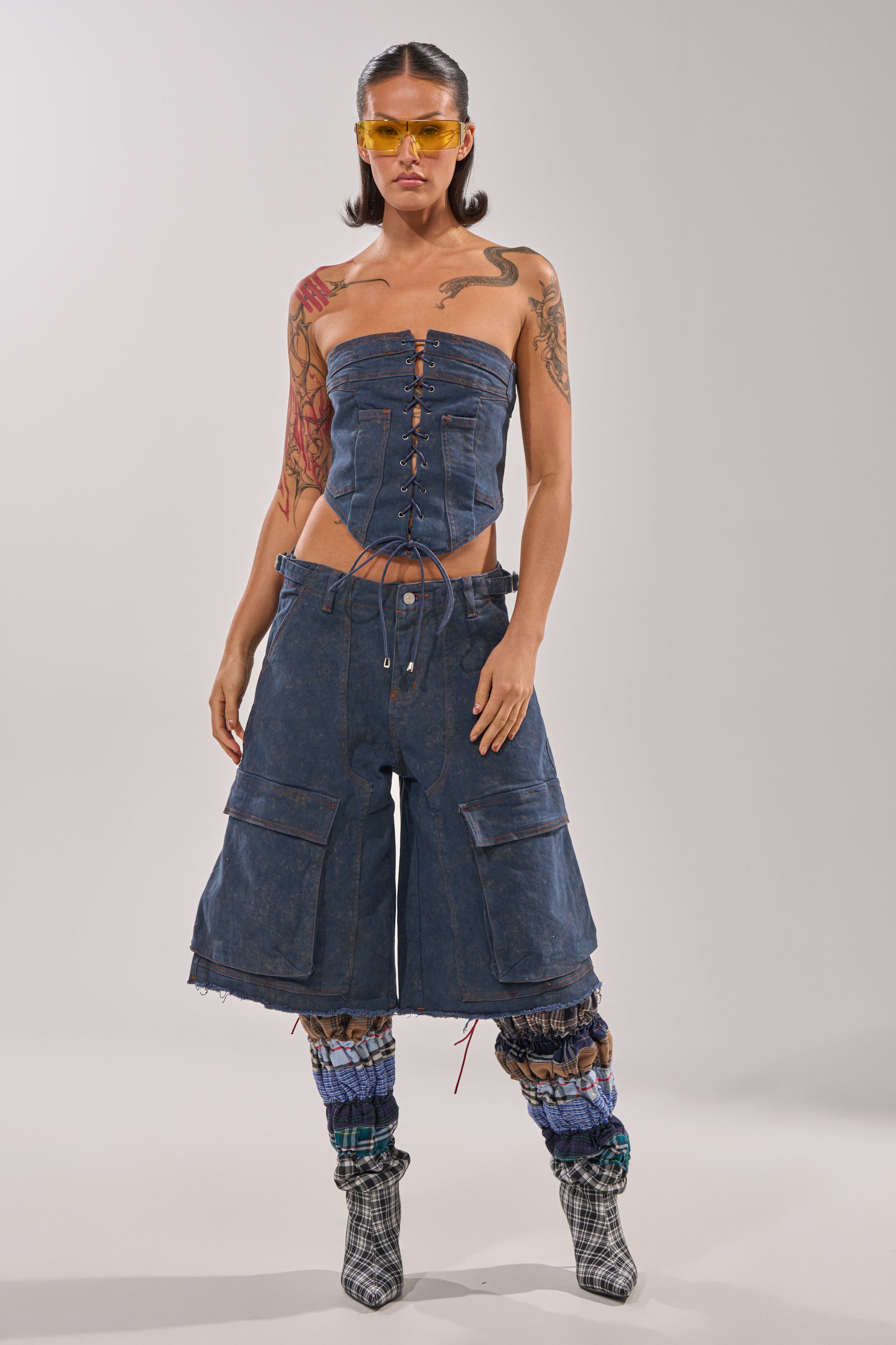 A woman in yellow sunglasses, a strapless lace-up denim corset, plaid leg warmers, checkered ankle boots, and BOLD BERMUDA SHORT dark-wash cargo shorts with a high-rise waist poses against a plain light gray background. 0