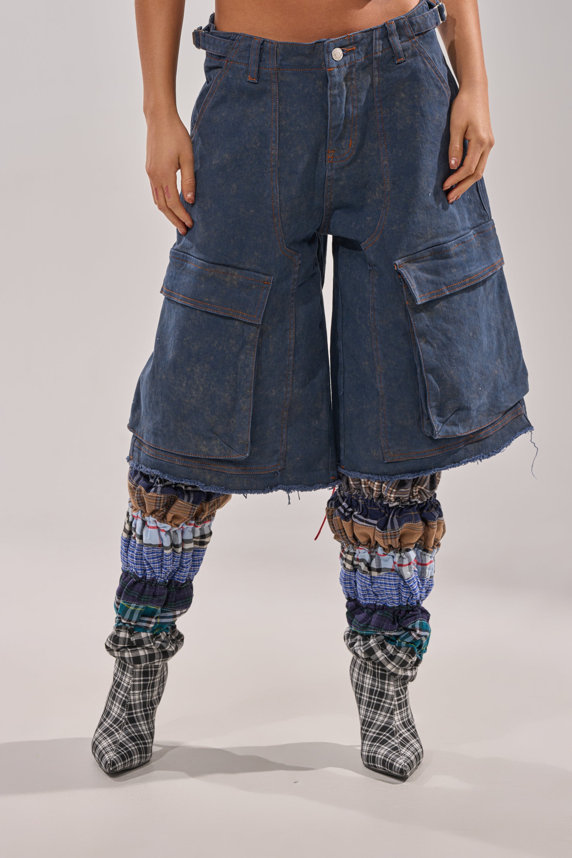 A person wearing the BOLD BERMUDA SHORT—high-rise, knee-length denim bermudas with cargo pockets—plus blue and brown patchwork leg warmers and black-and-white plaid heeled boots stands against a plain background. 1