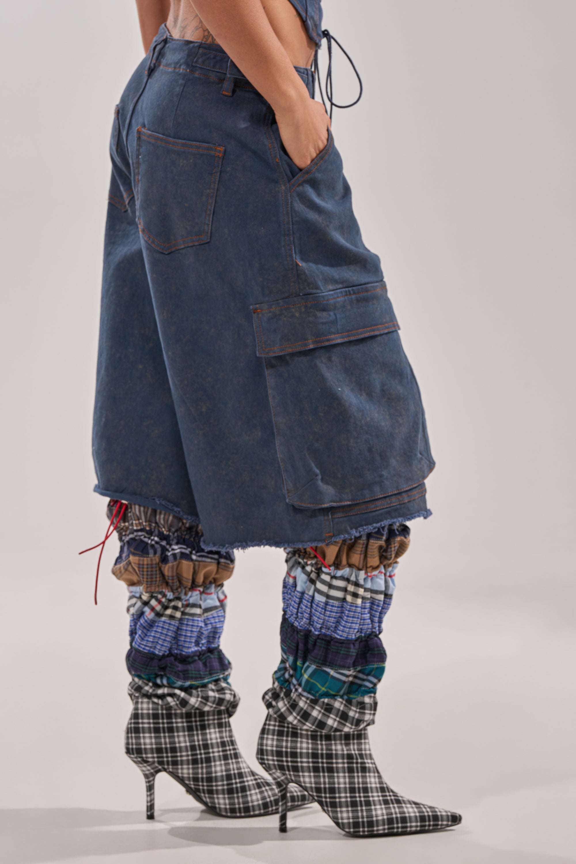 A person wearing BOLD BERMUDA SHORT denim bermuda shorts with a high-rise waist, fringe hems, patch and cargo pockets, plus colorful plaid leg warmers and black-and-white plaid high-heeled boots. Their hands are in their pockets. 2