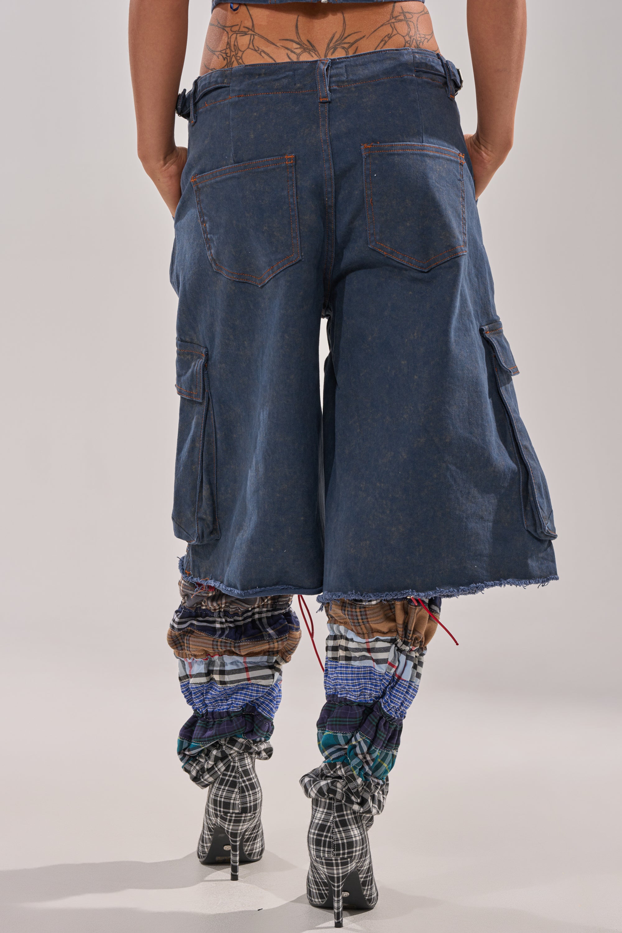 A person is shown from the waist down wearing the BOLD BERMUDA SHORT, featuring high-rise denim, large cargo pockets, and plaid fabric gathered at the calves. They pair it with high-heeled shoes against a plain, light background. 3