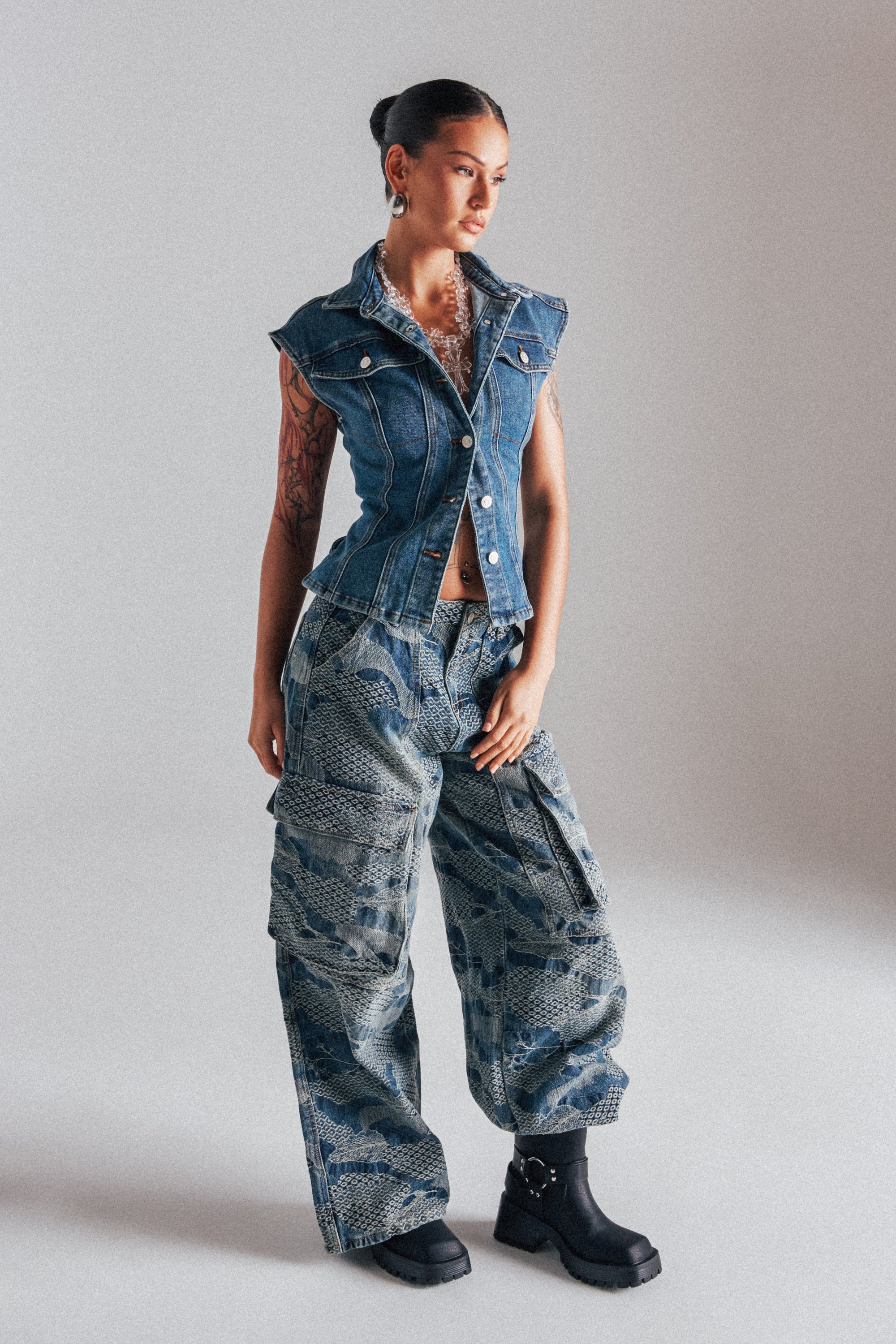 A woman with tattoos and tied-back hair stands in a studio wearing I THINK YOU WANNA KNOW DENIM CARGO PANT—a wide-leg blue denim pant—styled with a fitted sleeveless vest and chunky black boots. 1