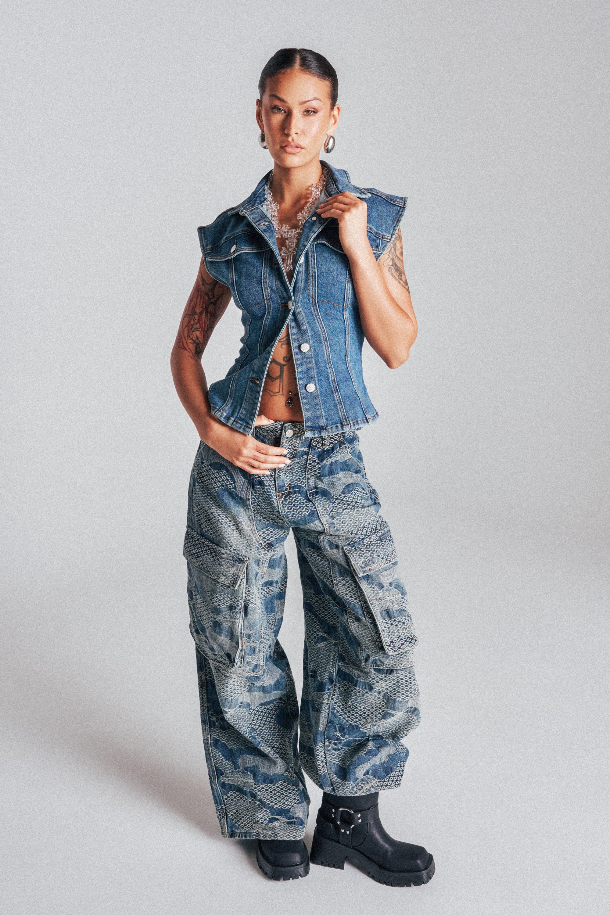 A woman stands confidently in the I THINK YOU WANNA KNOW DENIM CARGO PANT with a sleeveless denim vest, black boots, and large hoop earrings. Tattoos accent her arms as she poses against a plain light background. 2