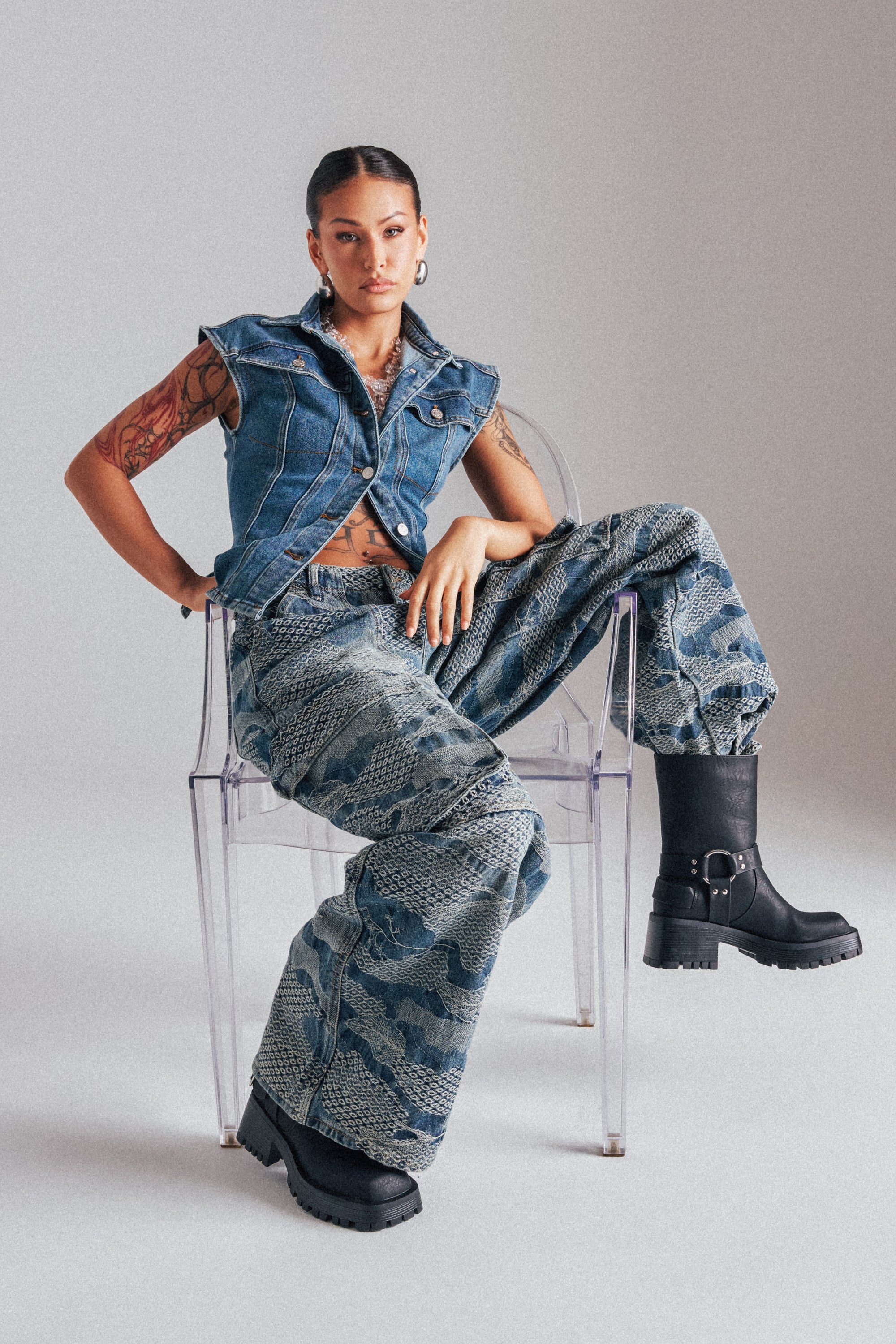 A tattooed woman sits confidently on a clear chair, wearing the I THINK YOU WANNA KNOW DENIM CARGO PANT with a sleeveless denim vest, black boots, slicked-back hair, and minimal accessories. 3