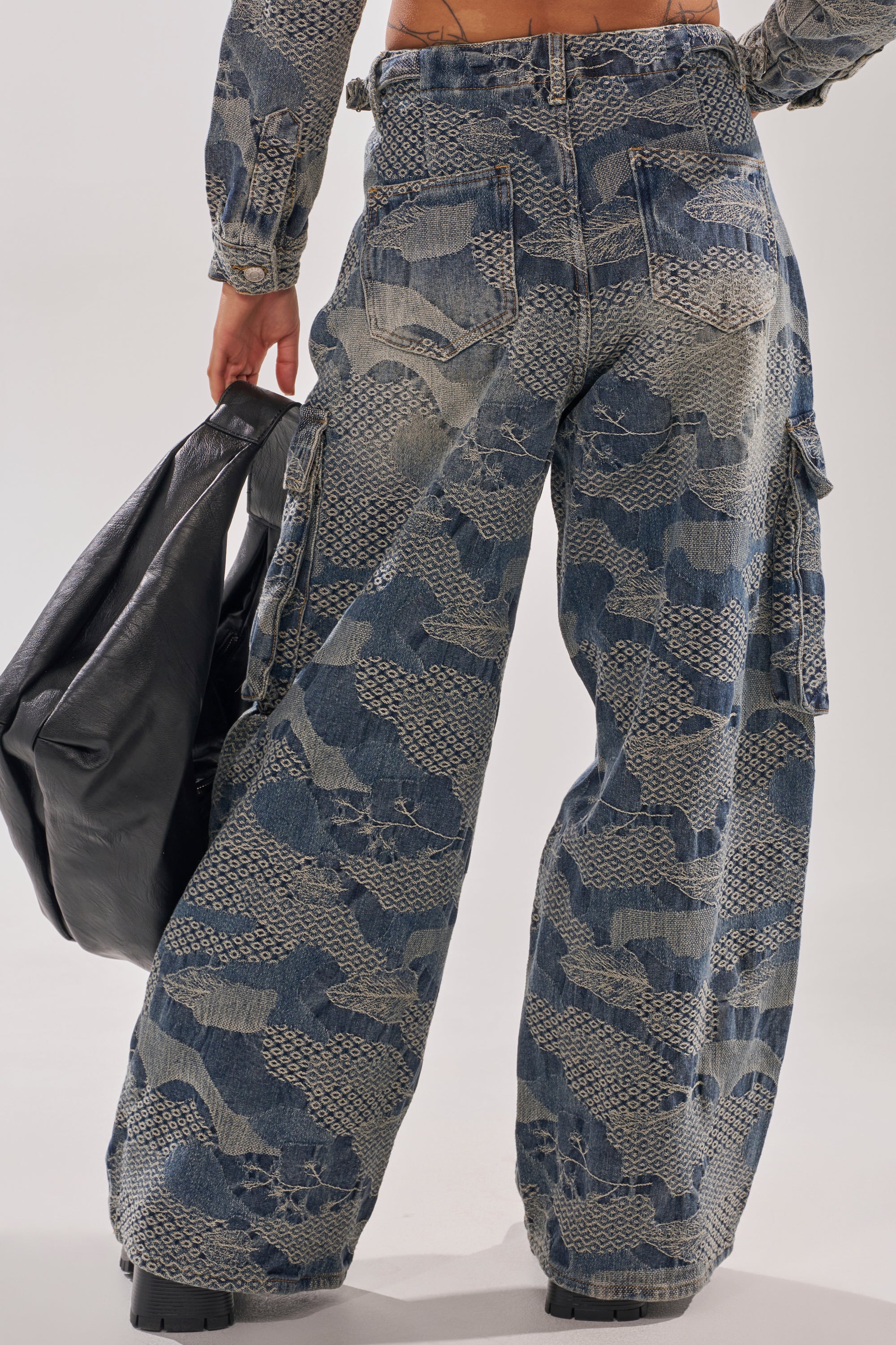 A person is seen from the back wearing I THINK YOU WANNA KNOW DENIM CARGO PANT, featuring a wide-leg fit, light snakeskin pattern, large cargo pockets, and paired with a big black bag. 7