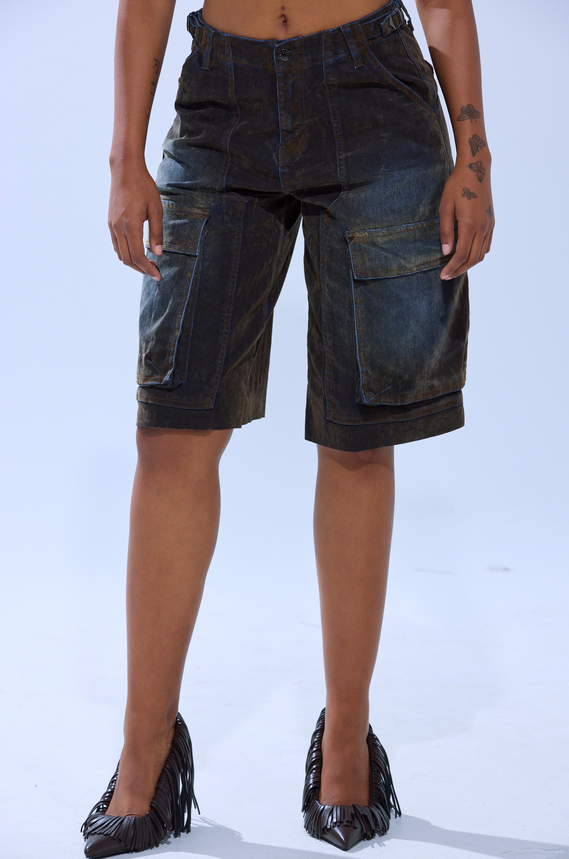 A person wears the BELLA VELVET DENIM JORT, a high-rise dark denim short with cargo pockets, paired with black pointed-toe fringe heels, standing against a plain light background.
