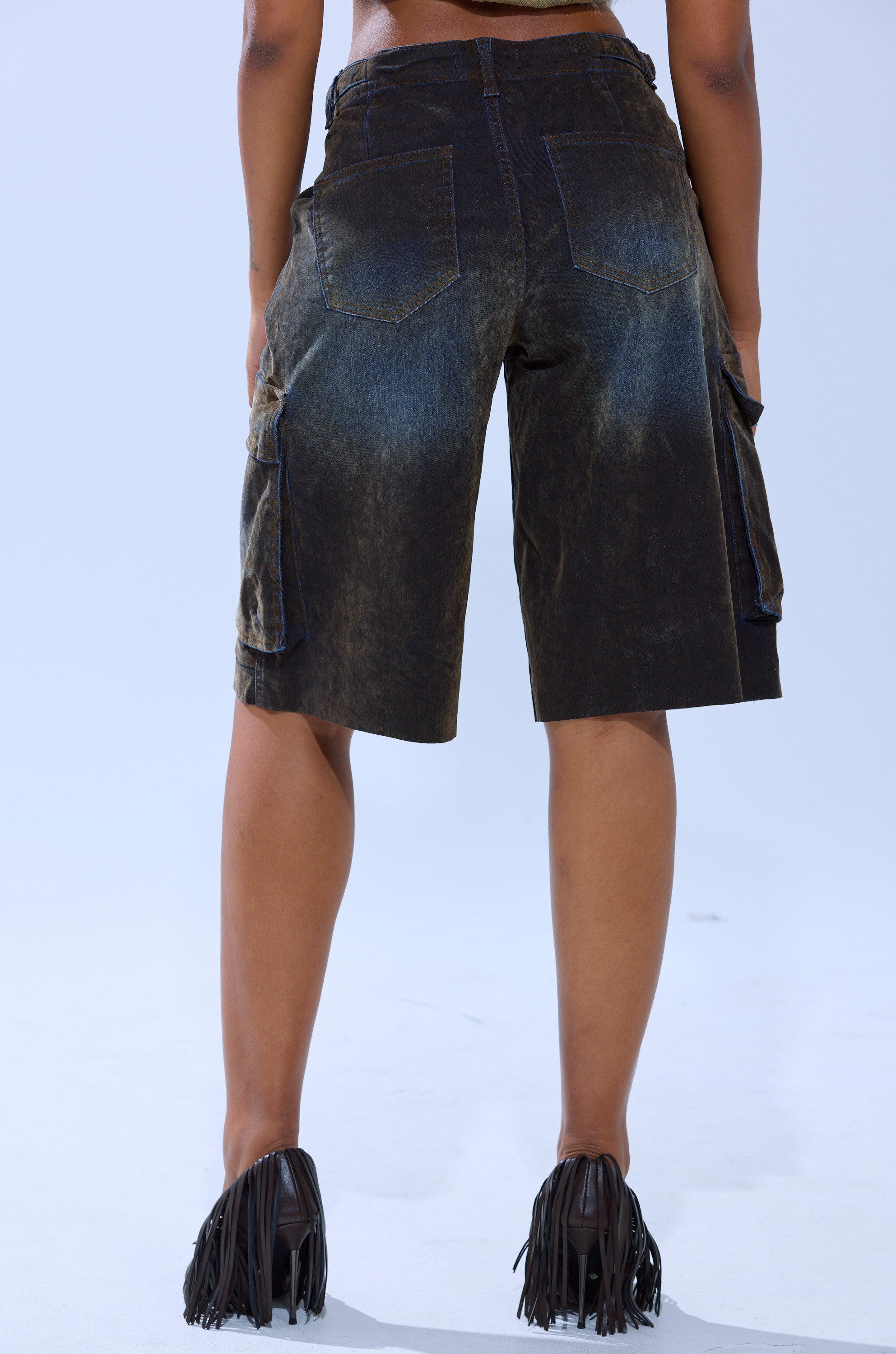A person faces away, wearing the BELLA VELVET DENIM JORT in faded black with cargo pockets and black fringe-detailed shoes, set against a plain light gray background.