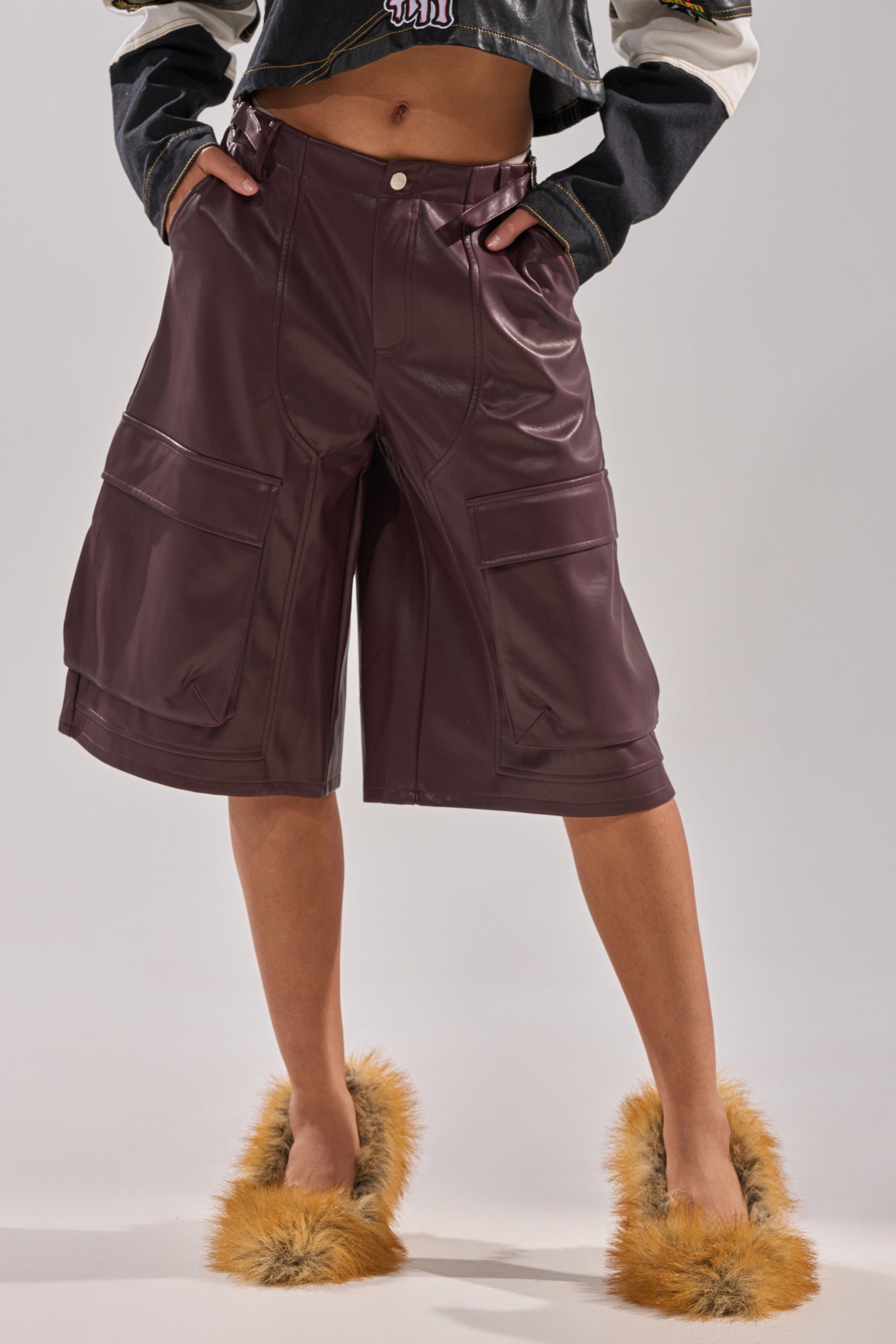 A person wears the OVER DONE FAUX LEATHER BERMUDA SHORT in dark brown with cargo pockets, paired with a cropped jacket and fluffy tan slippers. The photo is cropped to show only the lower half of their body.