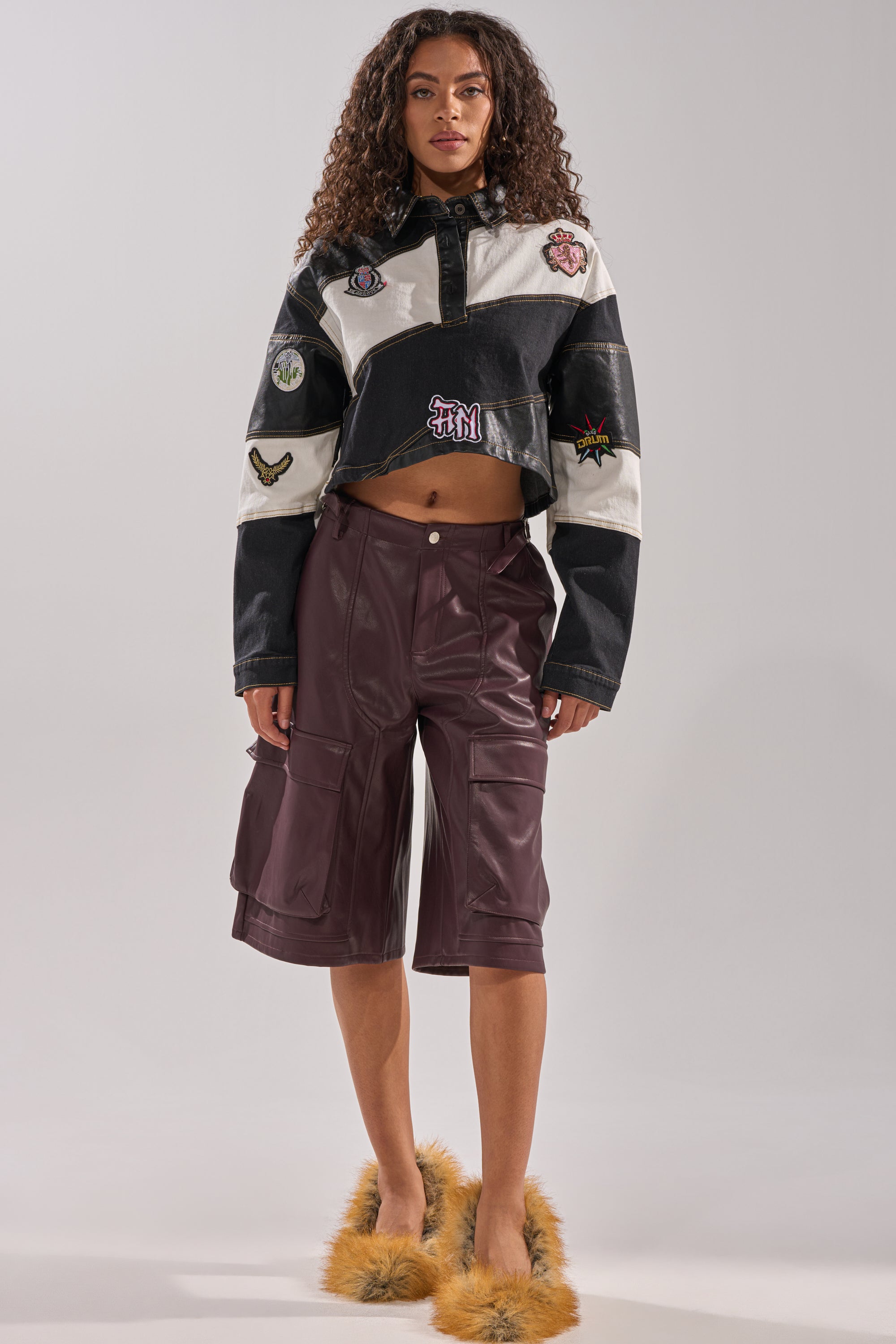 A woman with long curly hair and a neutral expression stands facing forward, wearing the OVER DONE FAUX LEATHER BERMUDA SHORT in burgundy, a cropped black-and-white patchwork jacket, and tan fuzzy slippers against a plain background.